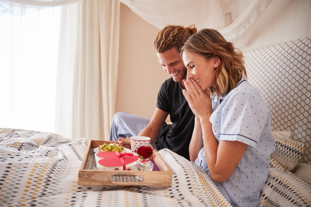 Millennial white man surprising his female partner with breakfast and gifts in bed, close up